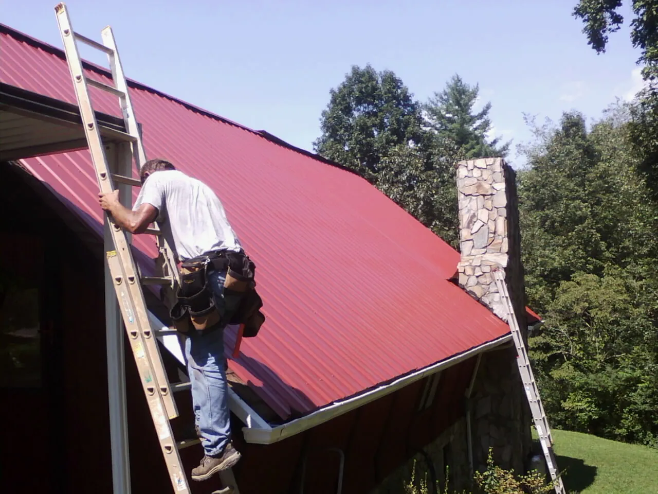 Roofer on a ladder beside a red metal roof and stone chimney — Soffit & Fascia Repair by Roofing Brothers in Orcas